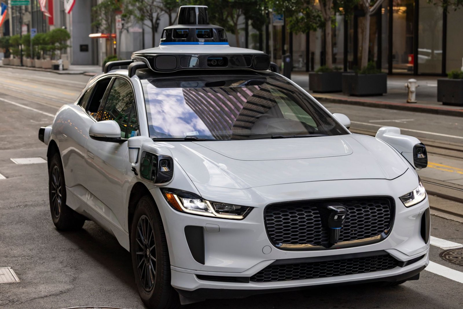 A white autonomous vehicle navigating a city street, reflecting urban architecture in daylight.