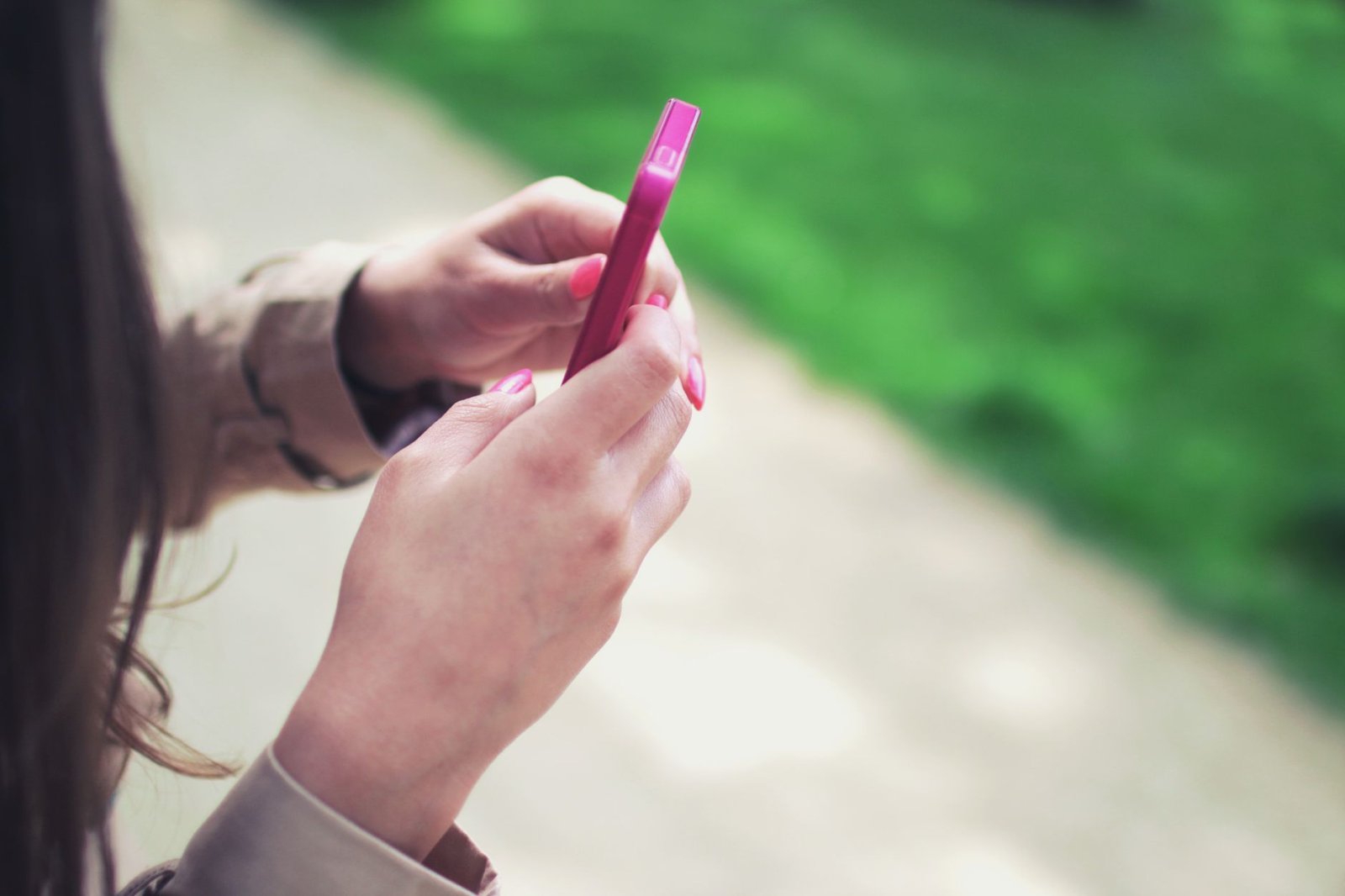 Woman holding a pink smartphone with both hands in a casual outdoor setting.