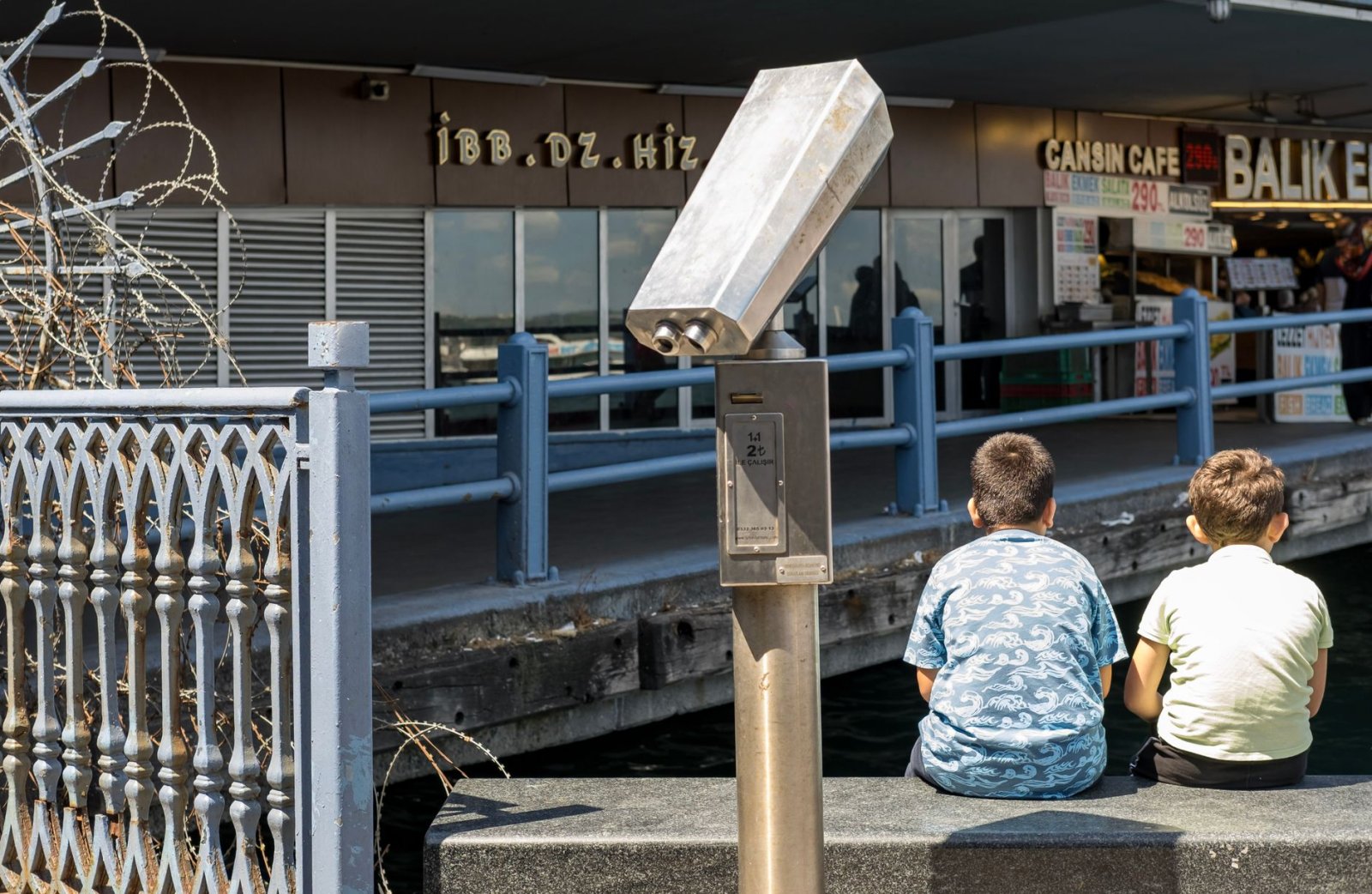 Two children sit by a waterfront, observing with binoculars. Sunny day scene.