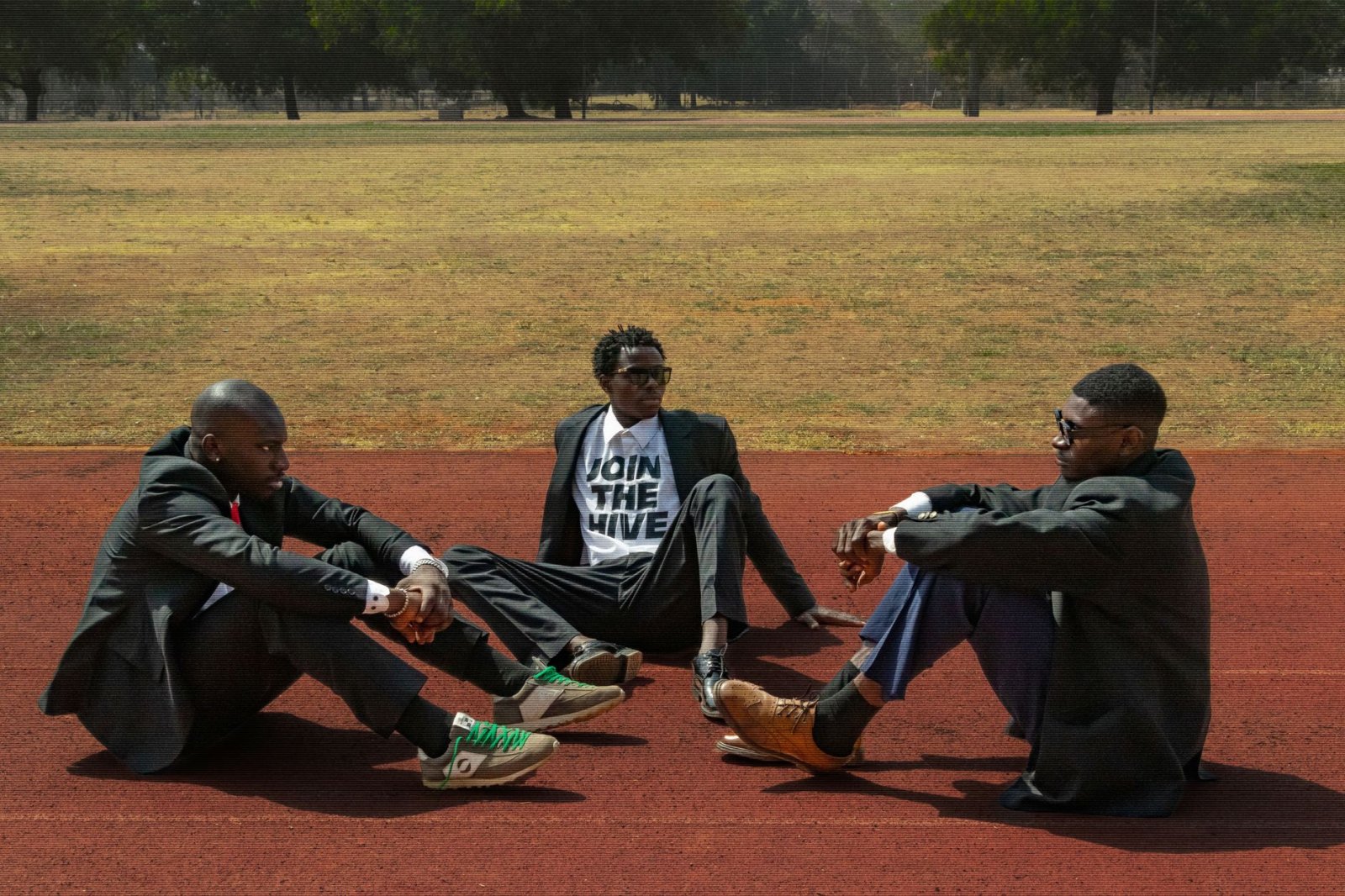 Three men in suits sit and converse on an outdoor track field, exuding modern style.